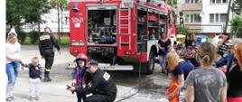 Zdjęcie przedstawia strażaka i dziecko w strażackim czerwonym hełmie. Strażak kuca i pokazuje dziecku jak lać wodę z prądownicy. W tle znajduje się budynek mieszkalny oraz czerwony ciężarowy samochód pożarniczy.