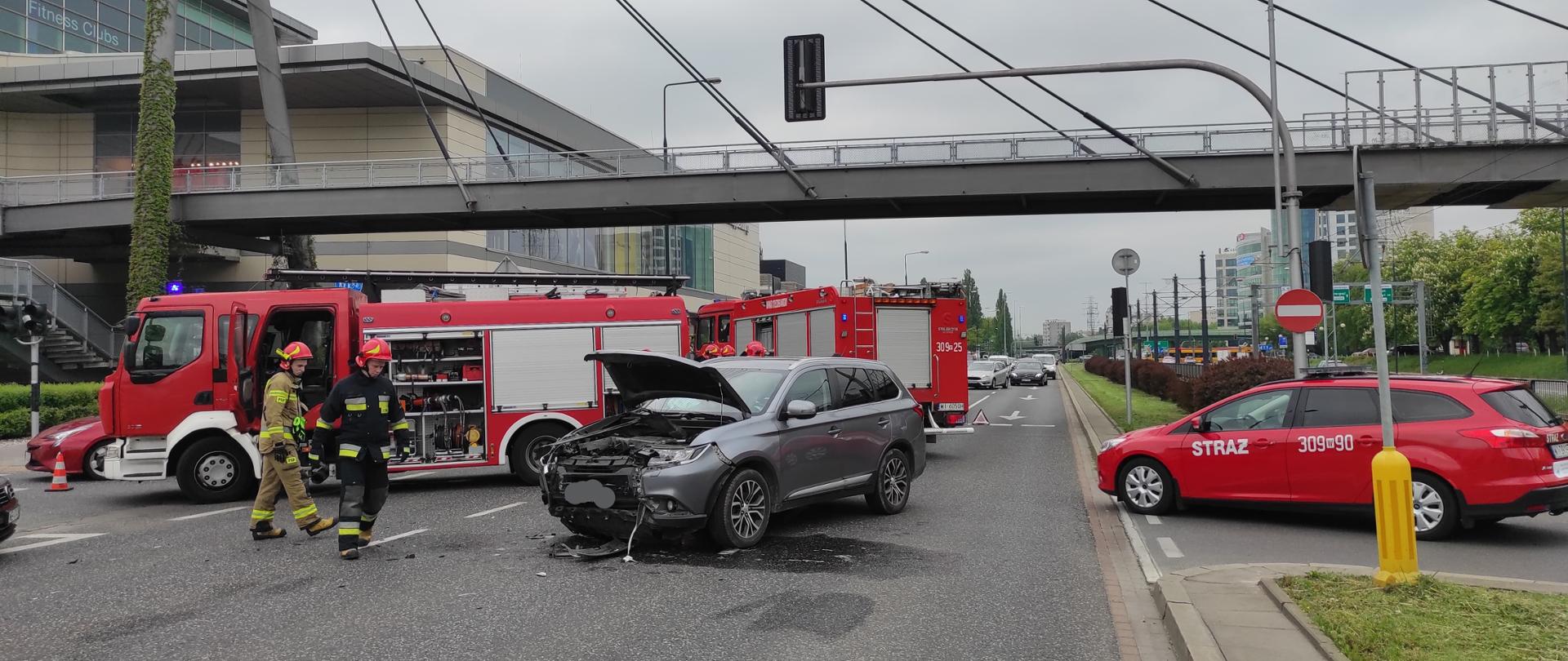 Na zdjęciu widoczny jest uszkodzony samochód osobowy, widoczne są trzy samochody straży pożarnej oraz pracujący strażacy. W tle widoczny jest budynek galerii handlowej