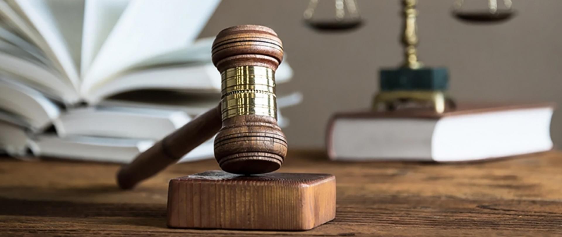 Młotek sędziego na podstawce, książki prawnicze i waga sprawiedliwości w tle.