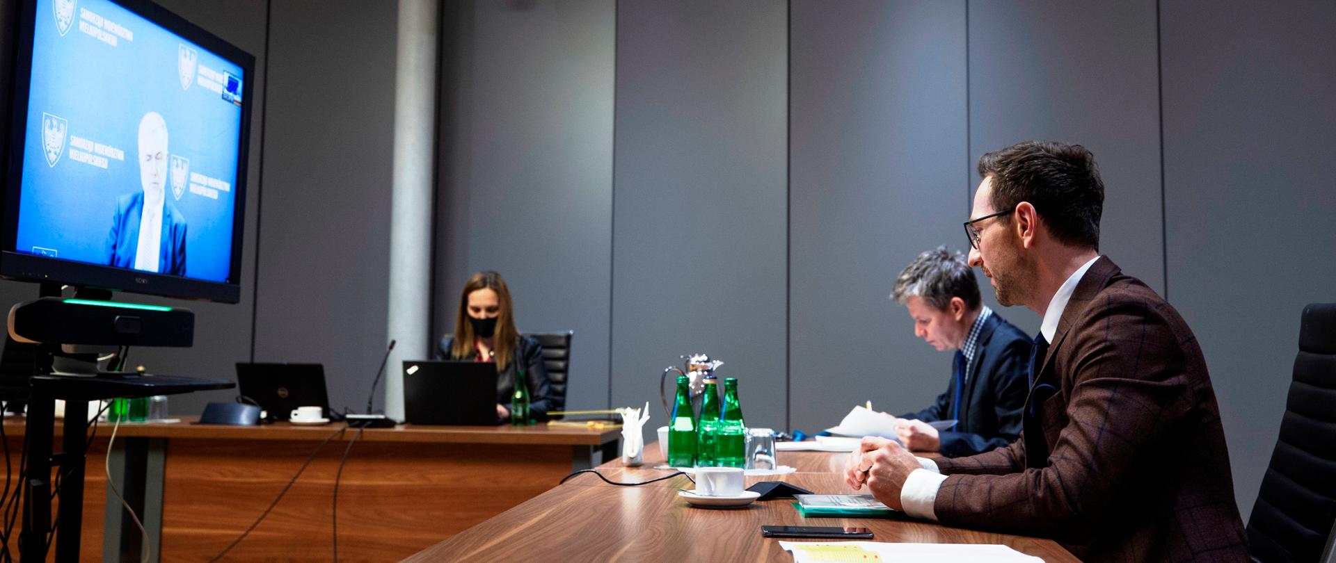 Na sali prze stołach siedzi dwóch mężczyzn i kobieta. Przed nimi ekran. Trwa zdalne spotkanie. Na pierwszym planie wiceminister Waldemar Buda.