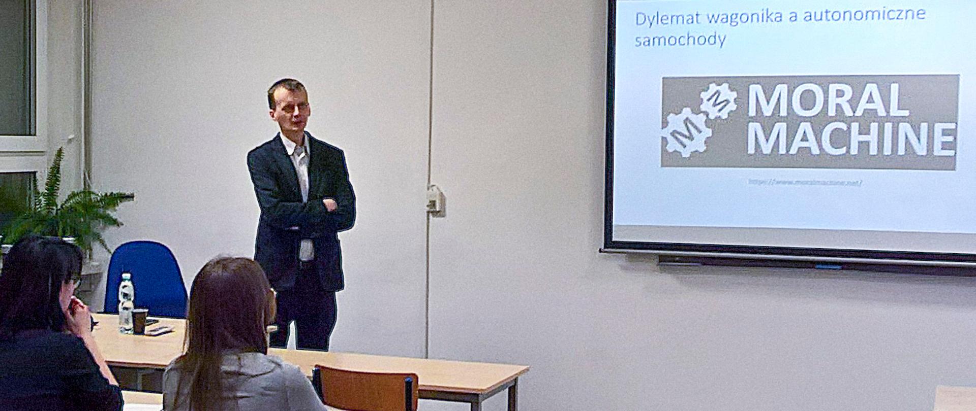 Sala lekcyjna, biurko, kilka ławek. Na ścianie przed nimi ekran, na nimi wyświetlony slajd prezentacji z tekstem: Moral machine, czyli dylemat wagonika a autonomiczne samochody. Przy jednej ławce siedzi czworo młodych ludzi, zwróconych w stronę mężczyzny, widzianych od tyłu. Przy biurku stoi mężczyzna, który patrzy na młodych ludzi. Młodzi ludzie patrzą na ekran