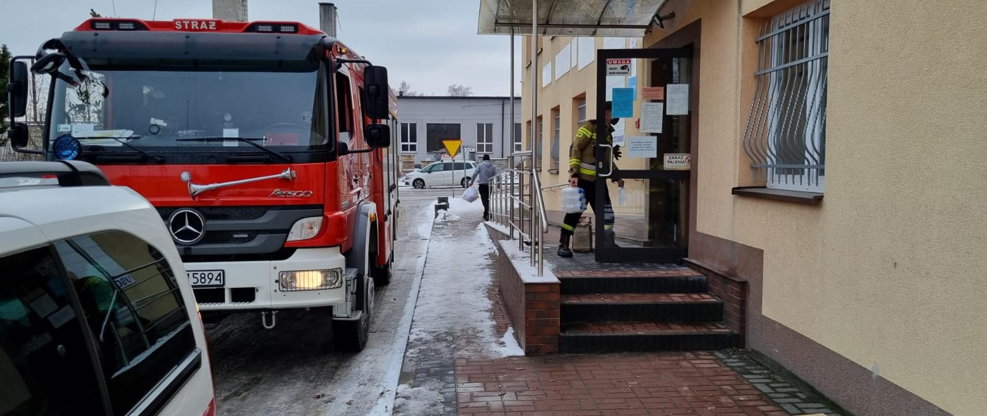 Zdjęcie przedstawia strażaka w umundurowaniu specjalnym, który wnosi środki ochrony indywidualnej do placówki medycznej, która stanowi także punkt szczepień przeciwko COVID-19. Zdjęcie zrobione przed placówką, przy samochodzie operacyjnym Komendy Powiatowej Państwowej Straży Pożarnej w Jędrzejowie oraz samochodzie ratowniczo-gaśniczym. Na zewnątrz zalega biała warstwa śniegu.