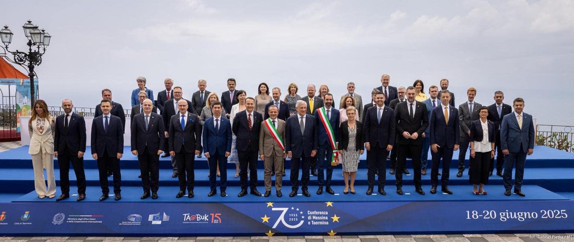 Duża grupa uczestników wydarzenia, ubranych w oficjalne stroje pozuje na tle pochmurnego nieba na niebieskim podeście z logotypem wydarzenia