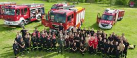 Grupa około 60 strażaków ustawiona jest do zdjęcia. Za nimi zaparkowane są wozy strażackie. W tle widać drzewa.