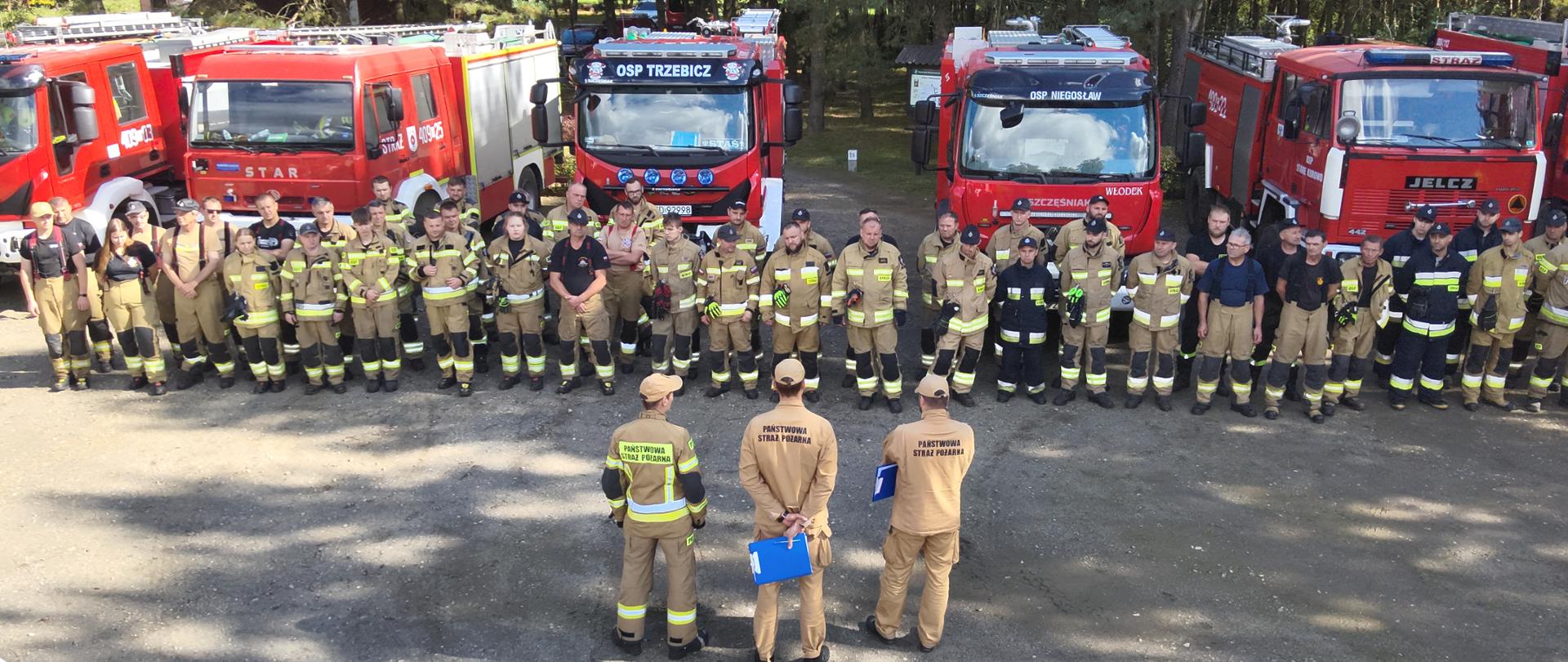 Fotografia przedstawia ujęcie z drona na plac przy leśnym centrum edukacyjnym. Na placu w rzędzie ustawione pojazdy pożarnicze biorące udział w ćwiczeniach przed pojazdami w dwuszeregu ustawieni ćwiczący strażacy przed nimi stoi dowódca ćwiczących pododdziałów. W tle drzewa.