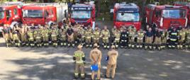Fotografia przedstawia ujęcie z drona na plac przy leśnym centrum edukacyjnym. Na placu w rzędzie ustawione pojazdy pożarnicze biorące udział w ćwiczeniach przed pojazdami w dwuszeregu ustawieni ćwiczący strażacy przed nimi stoi dowódca ćwiczących pododdziałów. W tle drzewa.