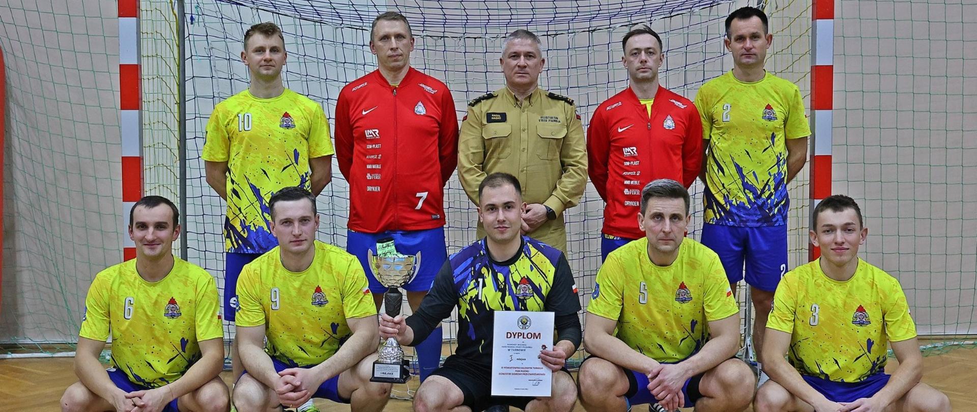 Wspólne zdjęcie drużyny z komendantem na tle bramki i boiska. Pierwszy rząd na kucąco, środkowy piłkarz trzyma w ręku puchar i dyplom