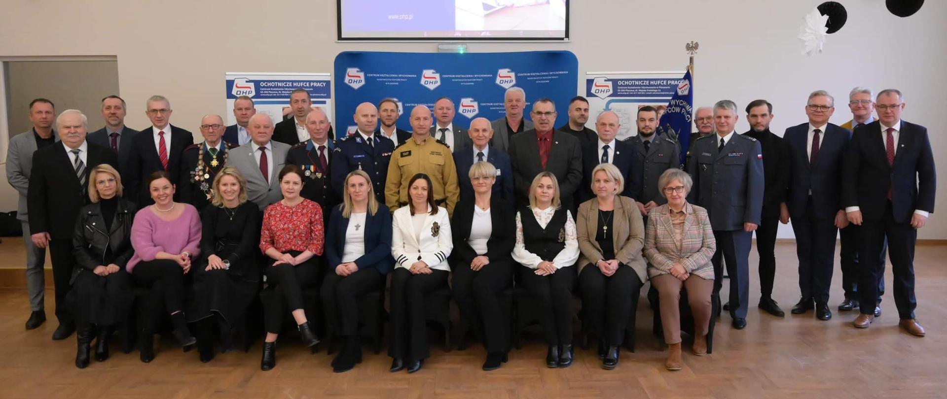 zdjęcie grupowe kobiet i mężczyzn ustawionych w trzech rządach w jasno świetlonej sali,, pierwszy rząd siedzi na krzesłach pośrodku drugiego rzędu widoczny mężczyzna w żółtym mundurze straży pożarnej, w tle znajdują się niebieskie banery Centrum Kształcenia i Wychowania Ochotniczych Hufców Pracy w Pleszewie