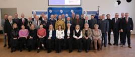 zdjęcie grupowe kobiet i mężczyzn ustawionych w trzech rządach w jasno świetlonej sali,, pierwszy rząd siedzi na krzesłach pośrodku drugiego rzędu widoczny mężczyzna w żółtym mundurze straży pożarnej, w tle znajdują się niebieskie banery Centrum Kształcenia i Wychowania Ochotniczych Hufców Pracy w Pleszewie