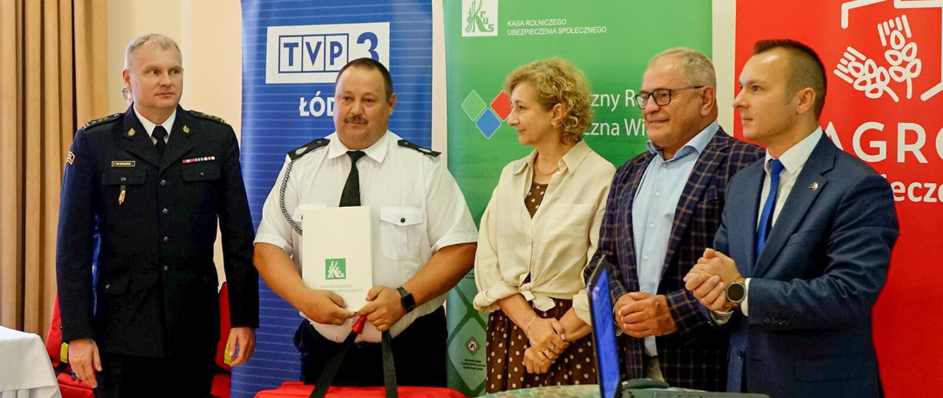 Pięciu dorosłych, w tym czterech mężczyzn i jedna kobieta, pozuje do zdjęcia. Mężczyzna w środku trzyma w rękach białą teczkę, a obok niego, na podłodze, leży czerwona torba medyczna. Tło stanowią banery z logo różnych organizacji, w tym TVP3 i KRUS.
