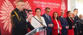 St. bryg. Rafał Świerczek przemawia przy mównicy, Obok niego stoją na tle ścianki z orłem białym, logo PSP oraz napisem Państwowa Straż Pożarna inni strażacy i osoby cywilne. 