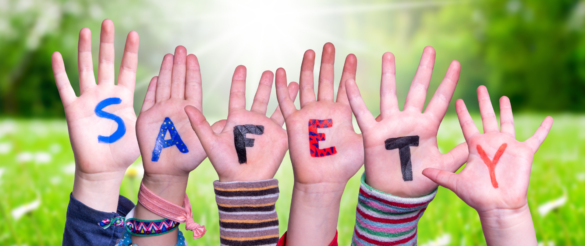 Baner przedstawia 6 dziecięcych rąk uniesionych do góry. Na każdej dłoni dziecka widnieje jedna litera - tworzą one łącznie napis SAFETY. W tle zielona polana z lasem i mocnym światłem na środku kadru.