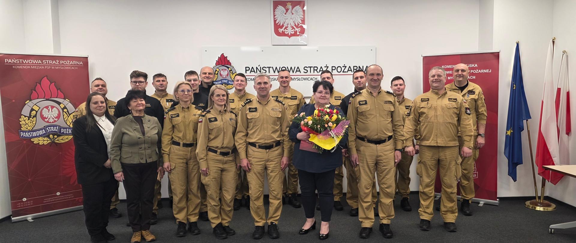 To zdjęcie stanowi oficjalne, pamiątkowe ujęcie grupowe, upamiętniające jubileusz 40-lecia pracy zawodowej Głównej Księgowej. Zostało wykonane w tej samej sali, co poprzednia fotografia, ukazując Jubilatkę w otoczeniu współpracowników i kierownictwa jednostki.
Kompozycja i uczestnicy
Na zdjęciu znajduje się liczna grupa osób (łącznie około 18 pracowników i funkcjonariuszy), ustawionych w dwóch rzędach:
W centrum: Jubilatka trzyma otrzymany bukiet kwiatów oraz czerwone etui. Po jej obu stronach stoją wyżsi rangą oficerowie Państwowej Straży Pożarnej w pełnym umundurowaniu służbowym.
Pozostali uczestnicy: Skład grupy to głównie strażacy w piaskowych mundurach oraz pracownicy cywilni. Obecność tak dużej grupy świadczy o silnych więziach wewnątrz komendy i wspólnym świętowaniu sukcesu koleżanki.
Symbolika i tło
Otoczenie podkreśla oficjalny i uroczysty charakter spotkania:
Symbole narodowe i formacyjne: W centralnym punkcie na ścianie wisi godło Polski (Orzeł Biały), a pod nim tablica z napisem "PAŃSTWOWA STRAŻ POŻARNA KM W MYSŁOWICACH" wraz z logo formacji. Z boku widoczne są flagi: Unii Europejskiej oraz Polski.
Oprawa graficzna: Po bokach grupy ustawiono pionowe roll-upy (banery) z logotypem Państwowej Straży Pożarnej, które domykają kompozycję i nadają zdjęciu charakteru oficjalnej dokumentacji z życia komendy.
Atmosfera
Zdjęcie jest statyczne i uporządkowane, typowe dla oficjalnych uroczystości państwowych. Wszyscy uczestnicy patrzą w stronę obiektywu, a na ich twarzach maluje się powaga wymieszana z życzliwością, co doskonale podsumowuje ten wyjątkowy moment docenienia czterech dekad profesjonalnej służby.