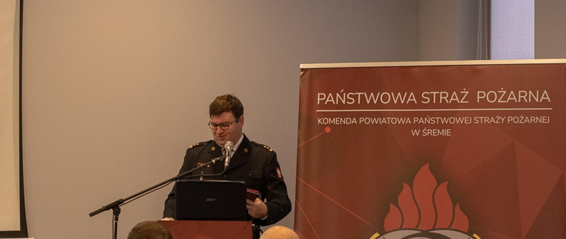 strażak stoi przed mównicą m za nim czerwony baner z logo państwowej straży pożarnej, przed nim siedzą ludzie
