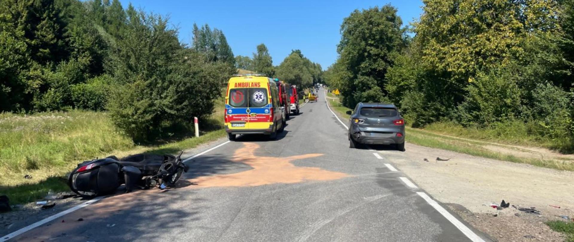 Zdjęcie przedstawia miejsce wypadku. Po lewej widać motocykl który leży na drodze i ma połamane sporo plastików. Od skutera na wprost w kierunku gdzie stoi karetka i dwa samochody gaśnicze straży pożarnej na drodze rozsypany jest sorbent.. Po prawej stronie na poboczu zaparkowany jest samochód który brał udział w wypadku z uszkodzonym lewym tyłem. Około 200 metrów od tego miejsca widać śmigłowiec LPR na drodze. 