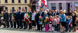 Plac 20 października w Śremie na płycie stoją dzieci z przedszkola z flagami i kwiatami obok opiekunowie dalej samorządowcy służby mundurowe 