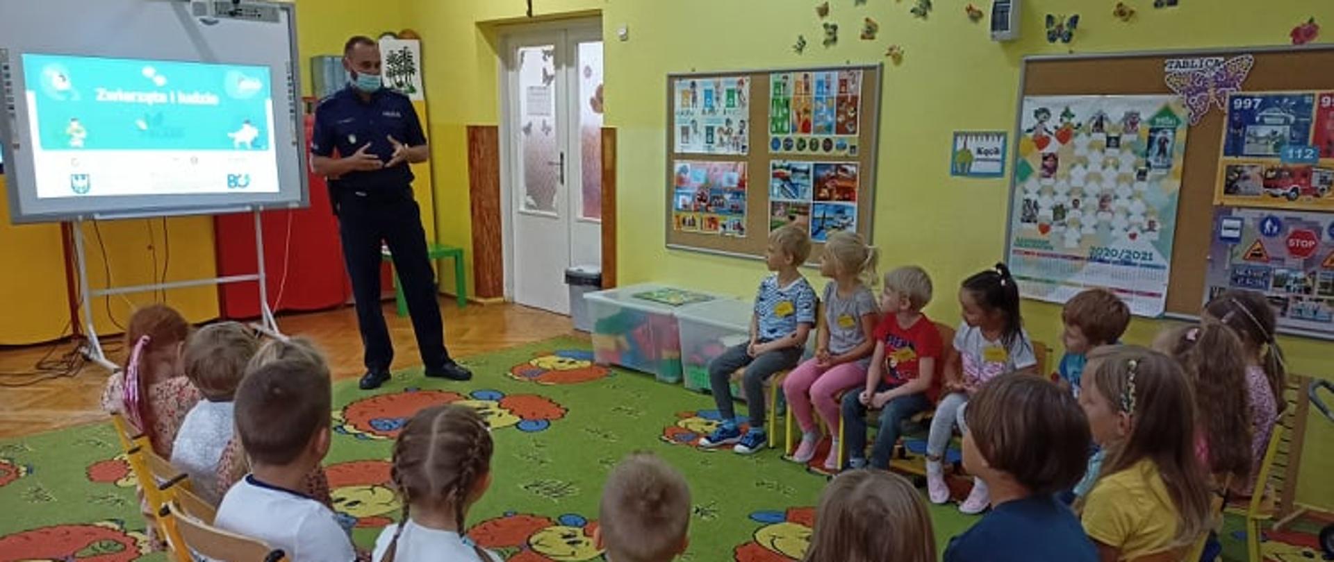 Grupa dzieci siedzi w kręgu i słucha policjanta prowadzącego warsztaty