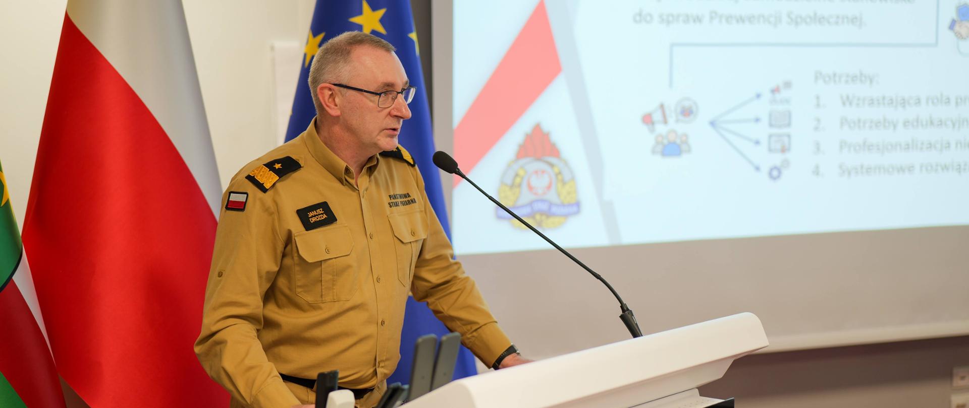 Mężczyzna w piaskowym mundurze służbowym straży pożarnej (nadbryg. Janusz Drozda) przemawia przy białej mównicy. Za nim widoczne są flagi: Polski oraz Unii Europejskiej, a w tle ekran z fragmentem prezentacji dotyczącym prewencji społecznej.