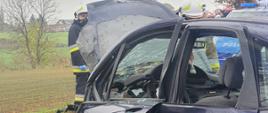 Widać uszkodzony samochód z lewym bokiem, podniesioną maską, w prawym rogu radiowóz Policji