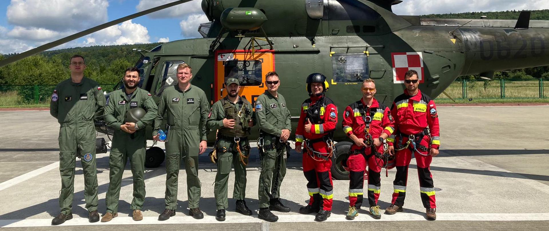 Zdjęcie przedstawia trzech strażaków wyposażonych w sprzęt ratownictwa wysokościowego wraz z pięcioma przedstawicielami Lotniczego Zespołu Poszukiwawczo Ratowniczego nr 8. W tle widoczny jest śmigłowiec W-3 "Sokół".