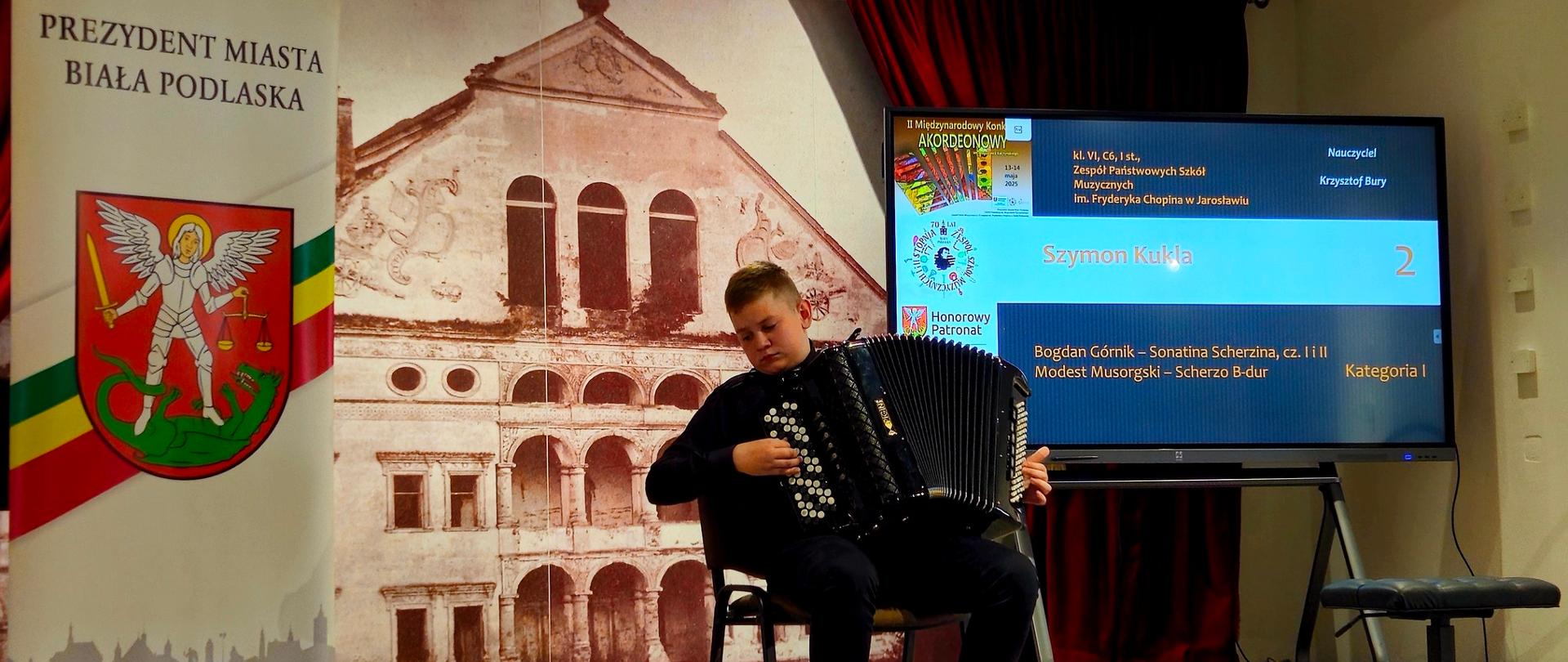 Na zdjęciu widoczny jest młody chłopiec grający na akordeonie, siedzący na czarnym krześle. Ubrany jest w ciemny strój, koszulę i spodnie, oraz brązowe buty. Po jego lewej stronie znajduje się wysoka, pionowa plansza z napisem „PREZYDENT MIASTA BIAŁA PODLASKA” oraz herbem miasta. W tle, za chłopcem, widać duży ekran telewizyjny, na którym wyświetlone są informacje dotyczące występu, w tym nazwisko „Szymon Kukla”, utwory, kategoria „Kategoria I” oraz nazwisko nauczyciela „Krzysztof Bory”. Na ekranie widoczne jest również logo „IV Międzynarodowego Konkursu Akordeonowego”. Z prawej strony ekranu znajduje się mały czarny stołek. W prawym górnym rogu zdjęcia widać fragment czerwonej zasłony, a w lewym górnym rogu ozdobny fragment ściany.