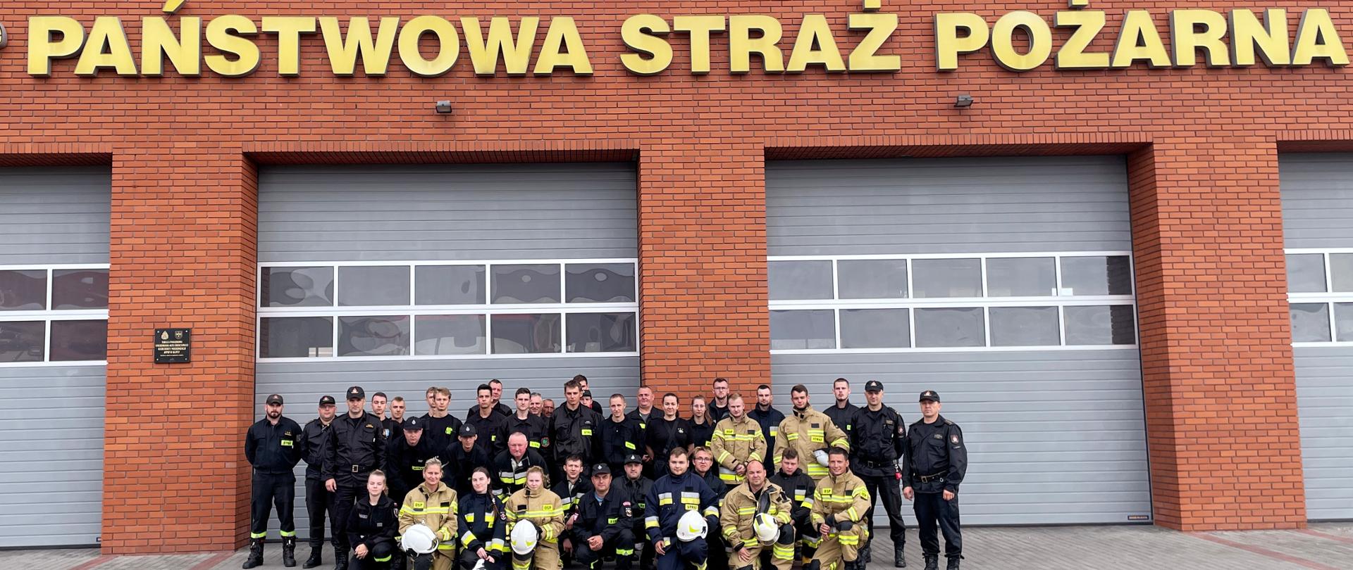 Zdjęcie grupowe uczestników kursu wraz z funkcjonariuszami KP PSP po zakończonym egzaminie. Zdjęcie na tle budynku z czerwonej cegły i napisem Państwowa Straż Pożarna