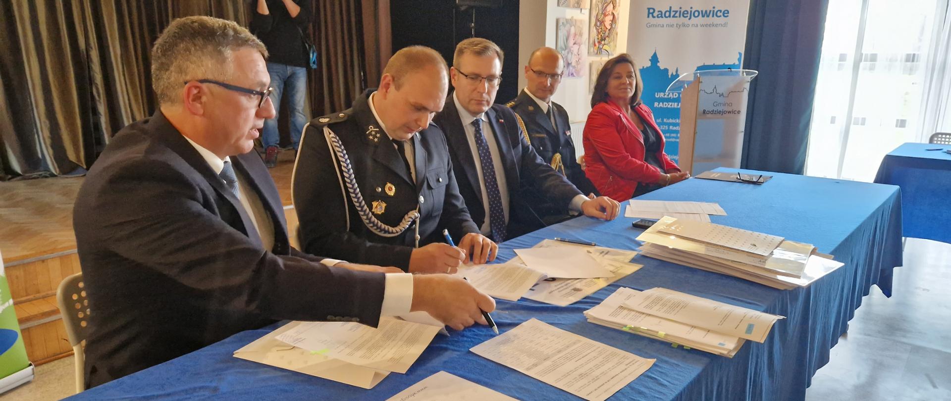 Strażak OSP podpisuje umowę. Po jego prawej stronie siedzi Prezes Zarządu Wojewódzkiego Funduszu Ochrony Środowiska i Gospodarki Wodnej w Warszawie Pan Marek Ryszka, po prawej kolejno : Sekretarz Stanu w Ministerstwie Aktywów Państwowych Pan Maciej Małecki, Komendant Powiatowy Państwowej Straży Pożarnej w Żyrardowie st. bryg. Mariusz Tymoszewicz oraz Wójt Gminy Radziejowice Pani Urszula Ciężka.