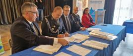 Strażak OSP podpisuje umowę. Po jego prawej stronie siedzi Prezes Zarządu Wojewódzkiego Funduszu Ochrony Środowiska i Gospodarki Wodnej w Warszawie Pan Marek Ryszka, po prawej kolejno : Sekretarz Stanu w Ministerstwie Aktywów Państwowych Pan Maciej Małecki, Komendant Powiatowy Państwowej Straży Pożarnej w Żyrardowie st. bryg. Mariusz Tymoszewicz oraz Wójt Gminy Radziejowice Pani Urszula Ciężka.
