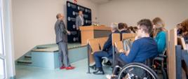 Zdjęcie z boku, minister Wieczorek stoi na tle granatowej ścianki z napisem Uniwersytet Kazimierza Wielkiego w Bydgoszczy i mówi do trzymanego w ręku mikrofonu, przed nim sala, w rzędach krzeseł siedzą ludzie, z boku mężczyzna na wózku inwalidzkim.