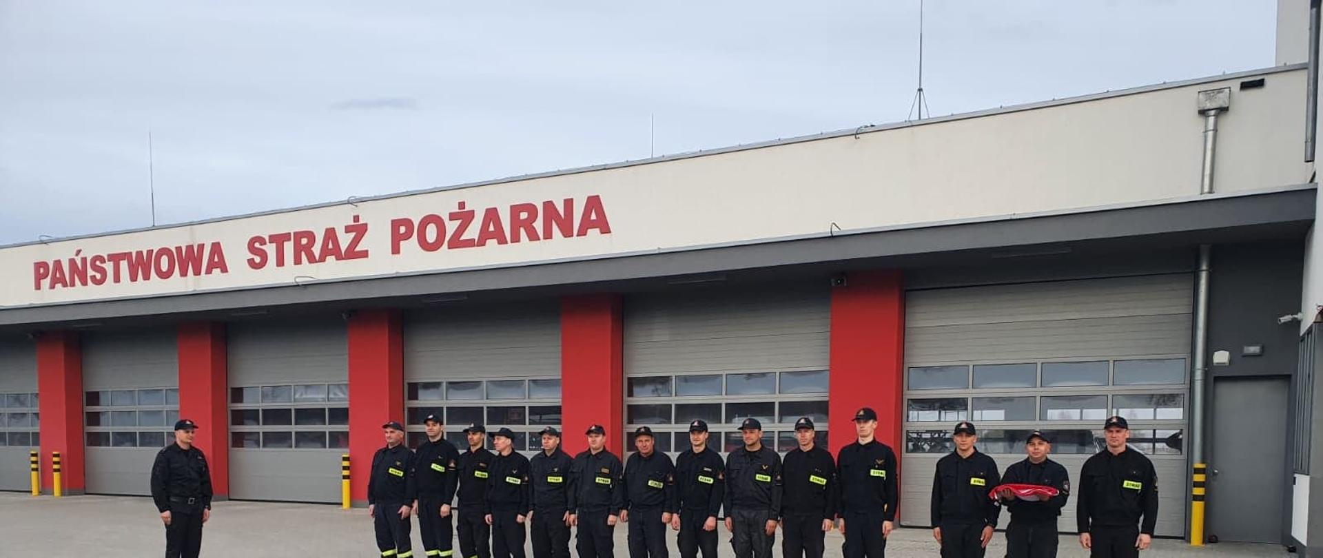Zdjęcie przedstawia piętnastu strażaków stojących przed budynkiem Komendy PSP