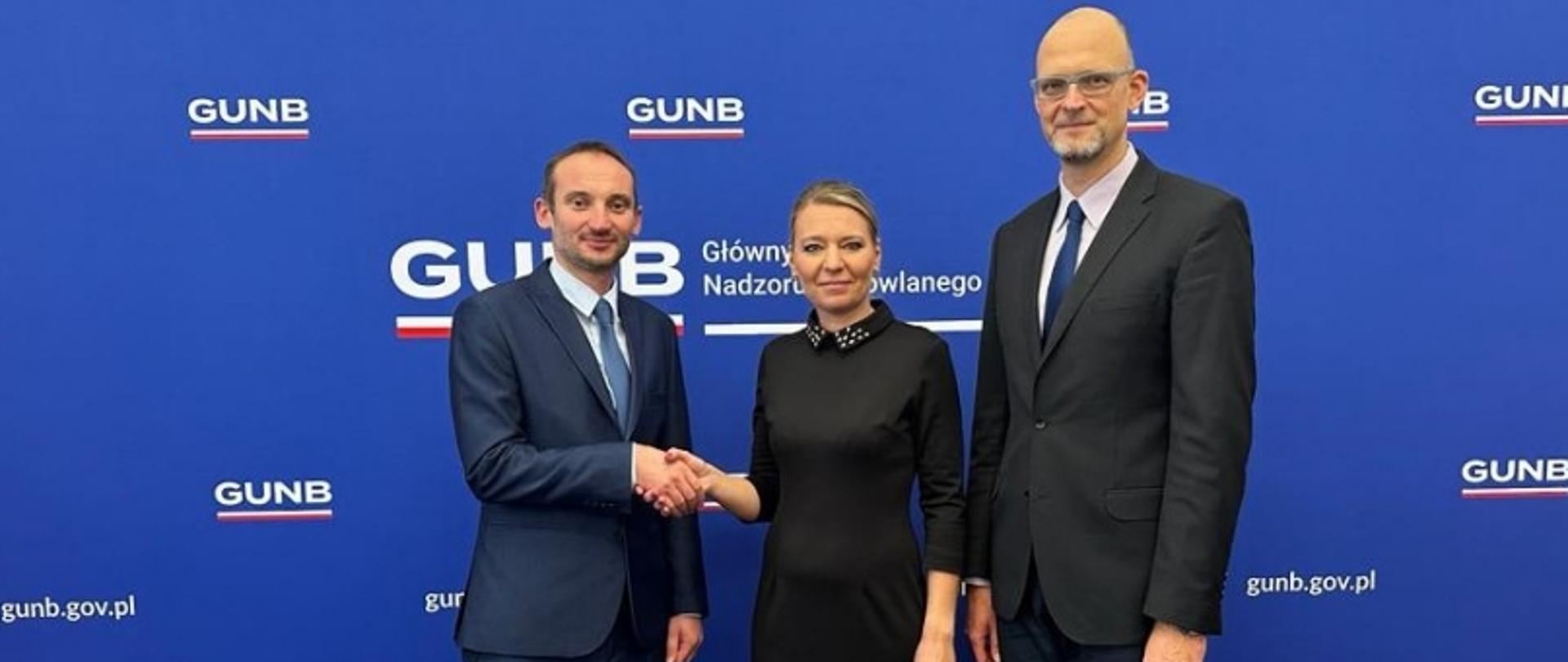 Three people standing in front of a blue backdrop with "GUNB Główny Urząd Nadzoru Budowlanego" and "gunb.gov.pl"; the person on the left is shaking hands with the person in the middle.