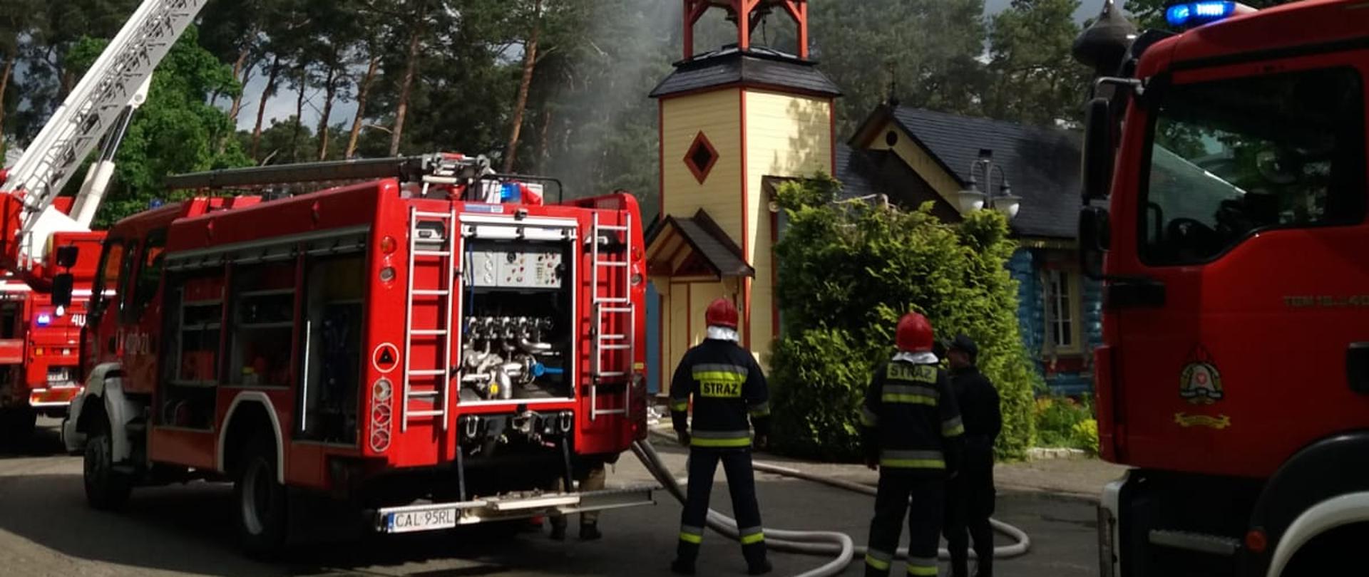 Na zdjęciu widać średni samochód ratowniczo-gaśniczy, ciężki samochód ratowniczo-gaśniczy oraz samochód specjalny z podnośnikiem hydraulicznym. Podnośnik jest rozstawiony oraz widać jak z niego podawana jest woda na budynek cerkwi. Z samochodu ratowniczo-gaśniczego widać rozwinięte linie wężowe, zarówno zasilające podnośnik jak i zasilające z hydrantu. Przy samochodzie widać stojących dwóch ratowników ubranych w ubrania specjalne i hełmy.