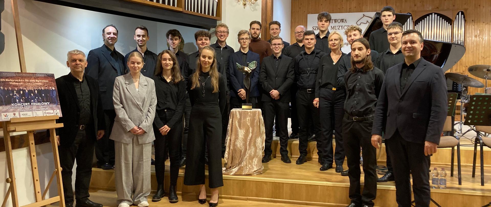 Międzynarodowa Orkiestra Jazzowa stoi na scenie w strojach galowych, w środku statuetka Nagrody Kulturalnej Śląska, po lewej plakat koncertu.