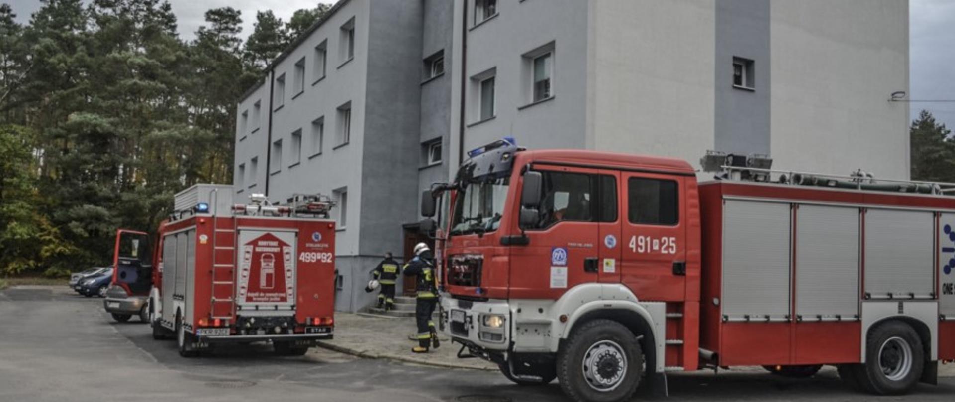 Przed budynkiem wielorodzinnym, stoją dwa samochody gaśnicze. Przed wejściem do obiektu widać dwóch strażaków w ubraniach bojowych. jeden z nich trzyma w ręku hełm. Nieopodal obiektu znajduje się las. 