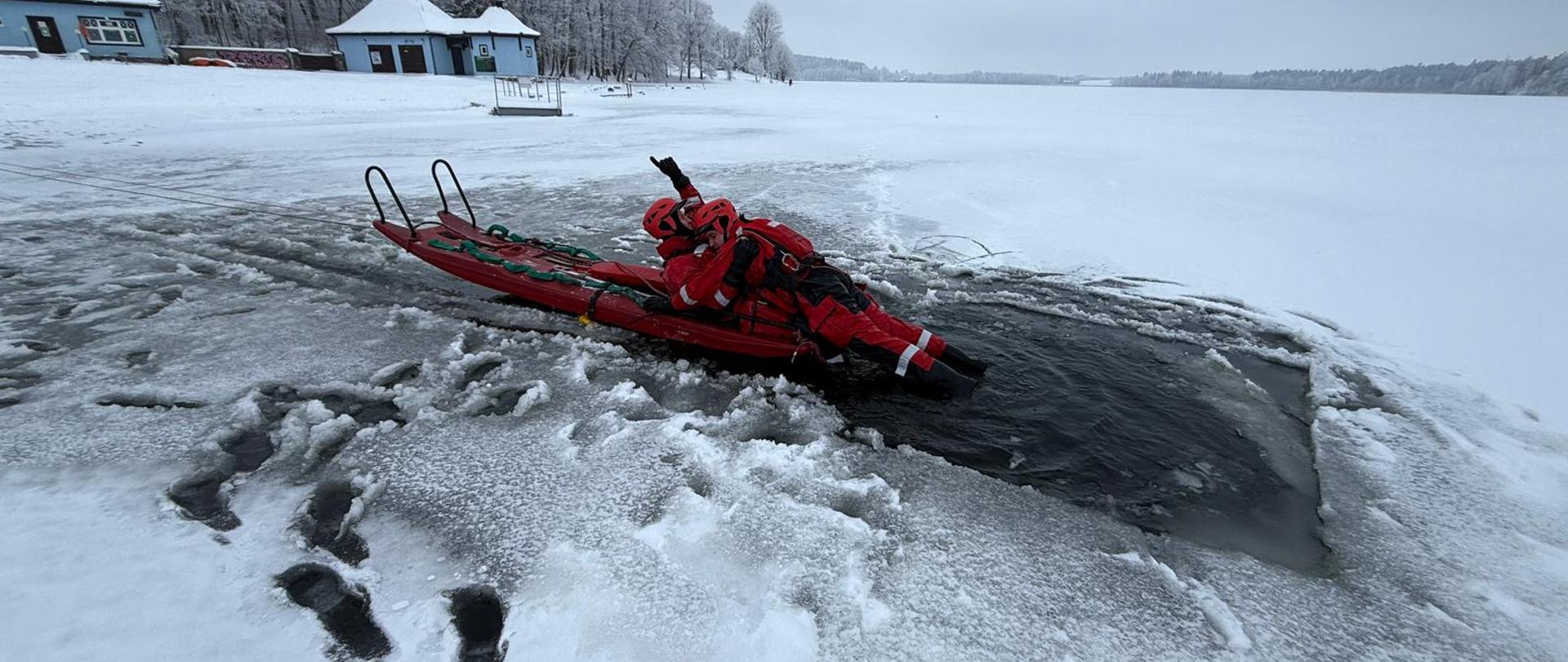 Strażacy w czerwonych ubraniach wypornościowych do pracy w wodzie ćwiczą podejmowanie osoby poszkodowanej, pod którą załamał się lód i wpadła do wody. Na zdjęciu strażacy na saniach lodowych. Zamarznięte jezioro w zimowej aurze.
