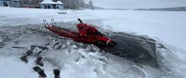 Strażacy w czerwonych ubraniach wypornościowych do pracy w wodzie ćwiczą podejmowanie osoby poszkodowanej, pod którą załamał się lód i wpadła do wody. Na zdjęciu strażacy na saniach lodowych. Zamarznięte jezioro w zimowej aurze.