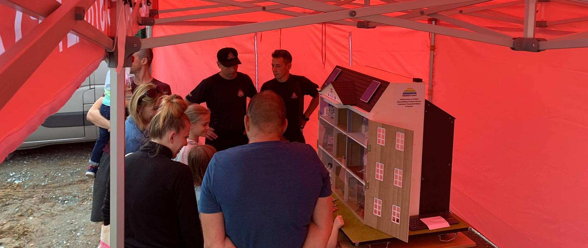 Pod namiotem komendy powiatowej znajduje się symulator zagrożeń budynku jednorodzinnego. Strażacy prowadzą pokazy z zakresu prewencji społecznej omawiając zagrożenia w obiektach mieszkalnych. Przed symulatorem stoi grupa słuchaczy w różnych kategoriach wiekowych. 