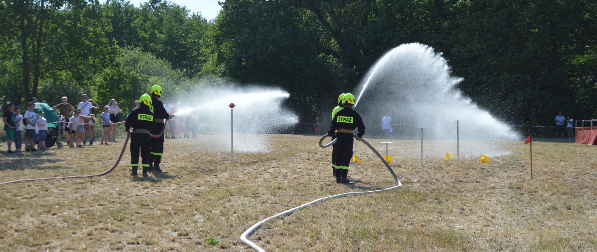 Zawody sportowo-pożarnicze w Jeziorach. Plac ćwiczeń. Trwa jedna z konkurencji zawodów - ćwiczenie bojowe. Końcowy etap ćwiczenia-obracanie tarczy przy pomocy strumienia wody oraz stracanie pachołków ustawionych na postumentach. zadania te wykonują dwie roty - 4 strażaków, którzy maja żółte hełmy. W tle drzewa i kibice.