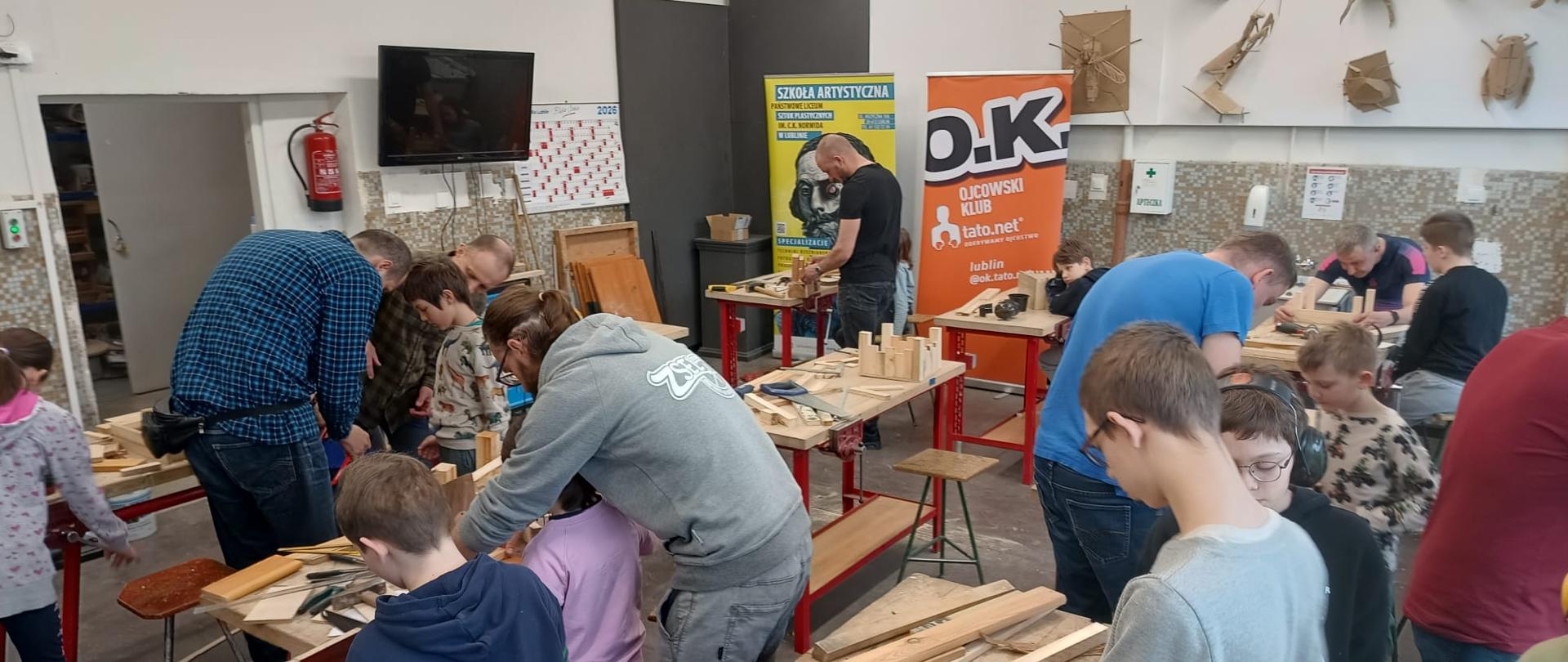 Warsztaty stolarskie dla dzieci i ojców w sali warsztatowej. Grupa osób pracuje przy stołach z drewnem, używając narzędzi ręcznych. W tle widoczne banery „Szkoła Artystyczna” oraz „Ojcowski Klub Tato.net”.