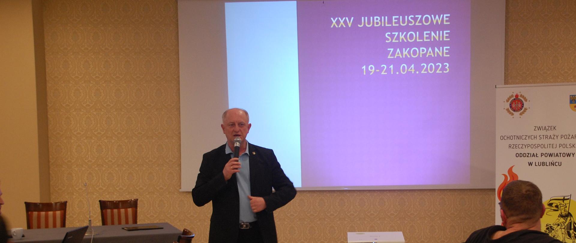 W centralnej części zdjęcia, na tle ekranu rzutnika multimedialnego, na którym wyświetlony jest slajd z napisem XXV jubileuszowe szkolenie Zakopane 19-21.04.2023, Prezes Oddziału Powiatowego Zarządu Związku Ochotniczych Straży Pożarnych Rzeczpospolitej Polskiej dh Krzysztof Strzoda przemawia do mikrofonu.