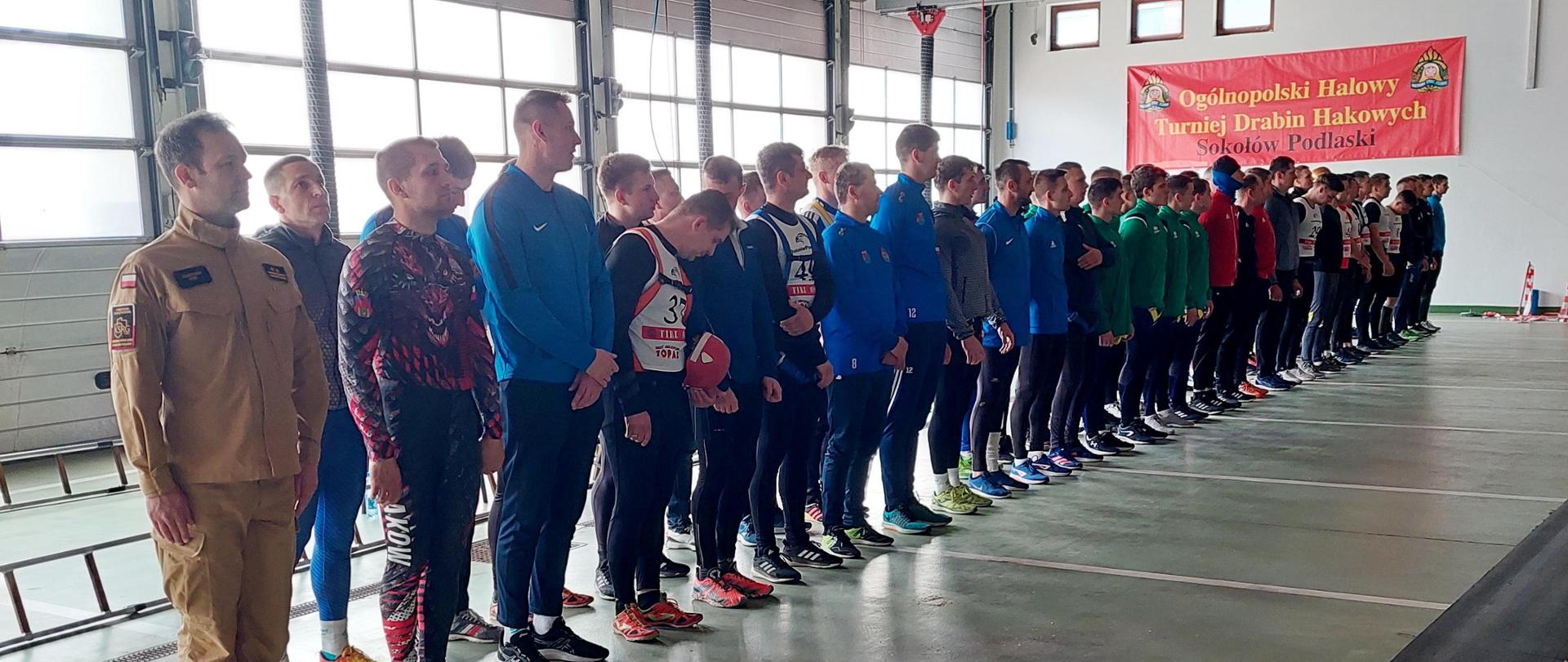 Zawodnicy stoją w dwurzędzie na sali gimnastycznej, na ścianie widnieje napis Ogólnopolski Halowy Turniej drabin Hakowych