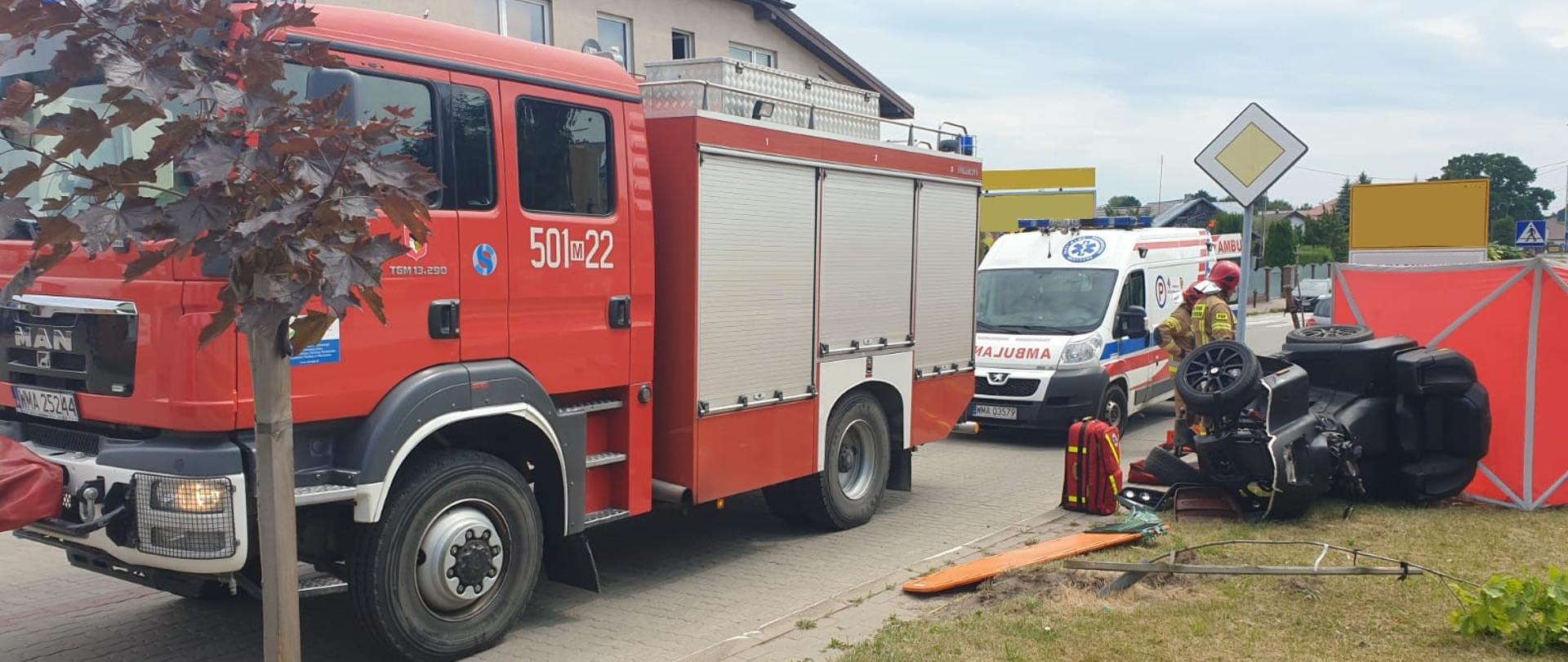 Quad po wypadku leży na boku na poboczu. Na drodze stoi zaparkowany samochód strażacki i karetka pogotowia.