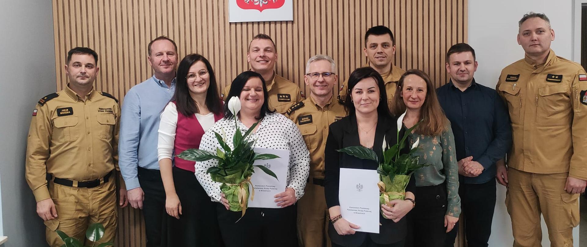 Załoga komendy wspólnie pozuje do zdjęcia z okazji Dnia Korpusu Służby Cywilnej. Okoliczności mają miejsce w gabinecie Komendanta Powiatowego Państwowej Straży Pożarnej w Brzezinach starszego brygadiera Mariusza Kaczmarskiego. Dwie Panie z korpusu cywilnego stoją w środku trzymają w ręku teczki gratulacyjne i kwiaty, które wręczył komendant. Na ścianie za pozującymi wisi godło Polski.