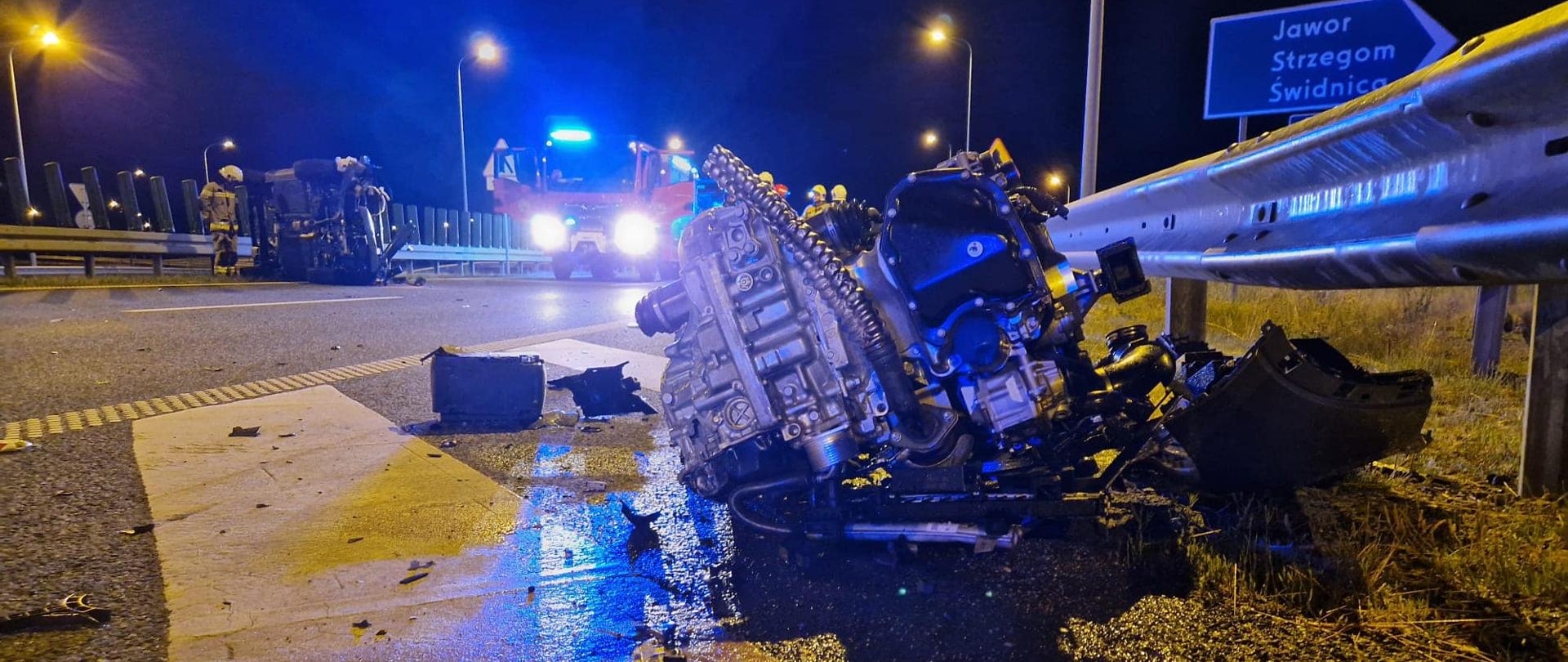 Obraz przedstawia silnik wyrwany z pojazdu wskutek wypadku. W tle samochód pożarniczy. Pora nocna