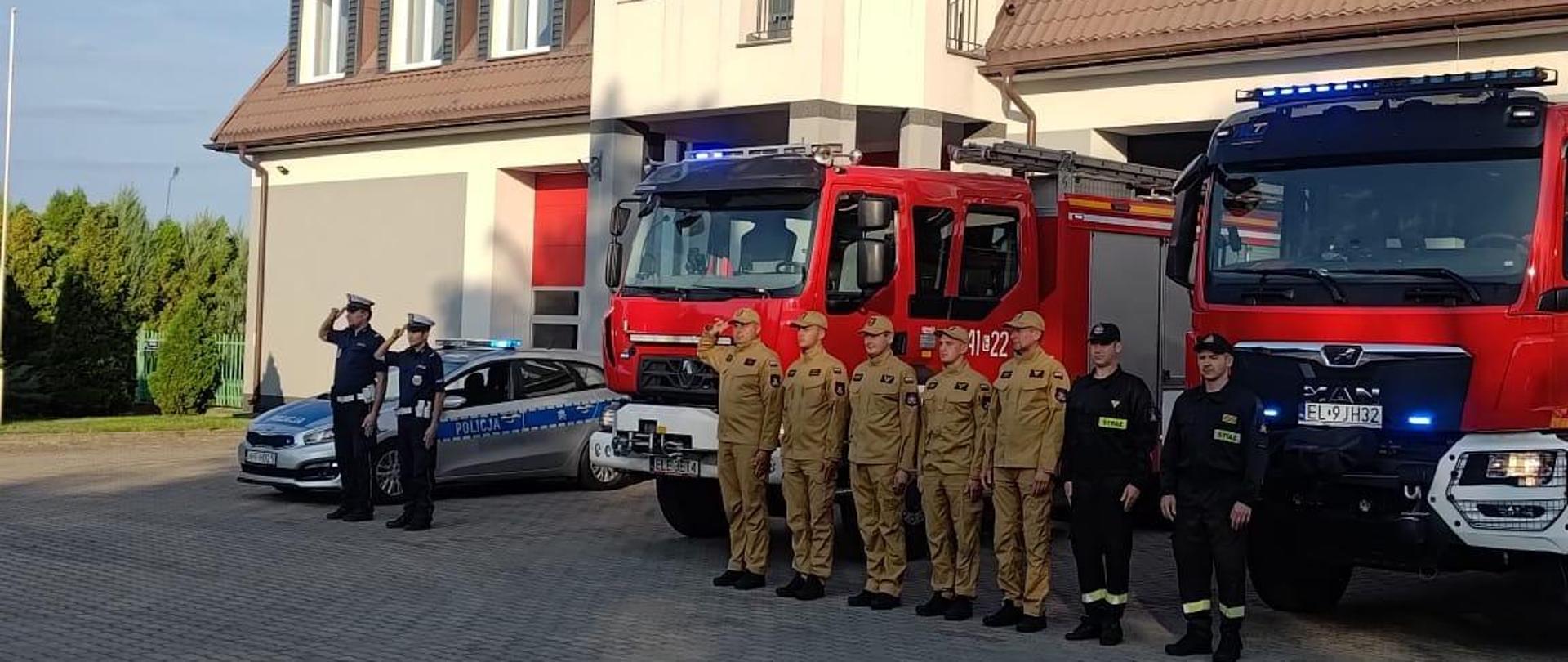 Na zdjęciu widać siedmiu strażaków stojących w jednym szeregu oraz dwa samochody strażackie na placu przed budynkiem Komendy Powiatowej PSP w Łęczycy. Dwóch strażaków ubranych jest w czarne stroje a pięciu w żółte. Wszyscy maja założone czapki z daszkiem. Za strażakami ustawione są dwa czerwone samochody strażackie, które mają włączone światła mijania oraz niebieskie światła do jazdy alarmowej. Obok stoją dwaj policjanci ubrani w niebieskie mundury i czapki. Obok zaparkowany jest radiowóz policyjny, który ma włączone niebieskie światła do jazdy alarmowej. Jest słonecznie.