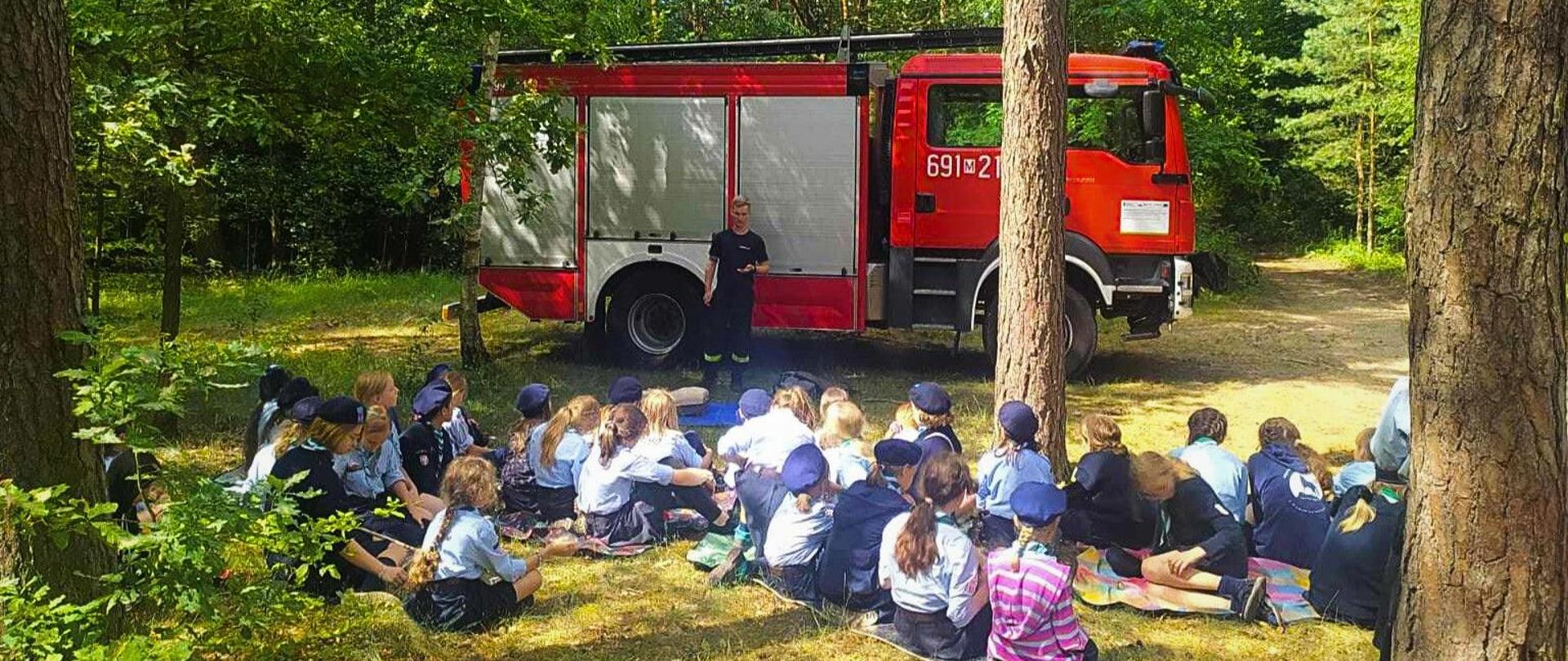 Strażak stoi przed samochodem ratowniczo-gaśniczym i prowadzi prelekcję dla siedzących przed nim na trawie harcerzy