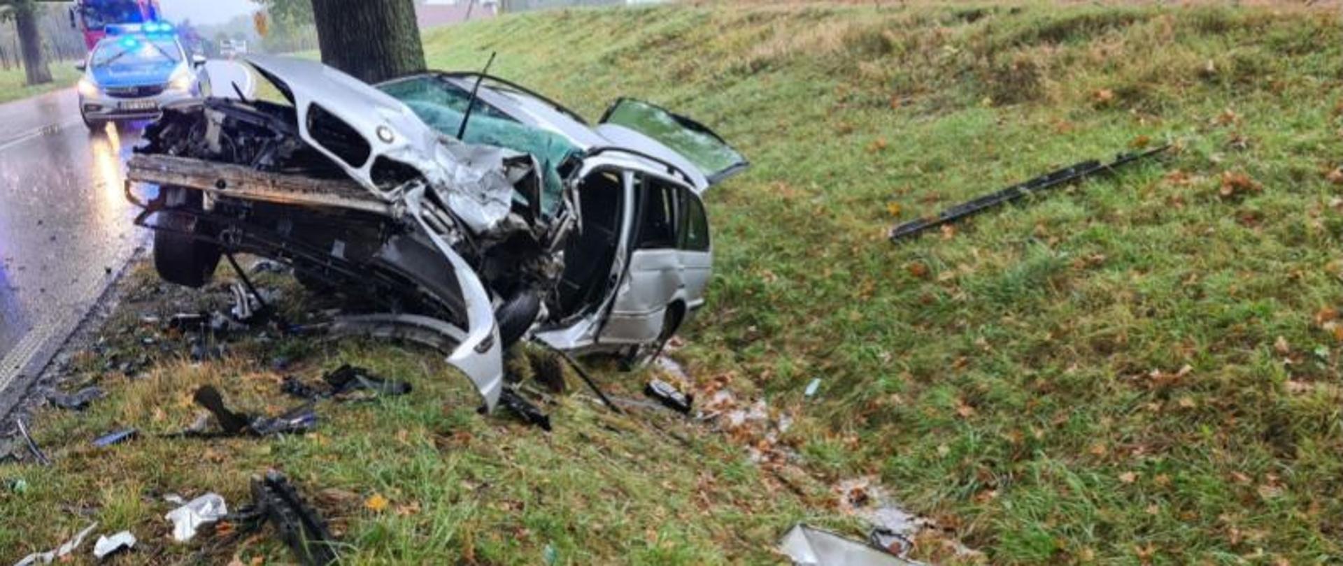 Zdjęcie przedstawia rozbity pojazd koloru srebrnego, który zatrzymał się na drzewie na poboczu drogi. Dookoła porozrzucane są elementy karoserii samochodu. W tle widać radiowóz i pojazd straży pożarnej.
