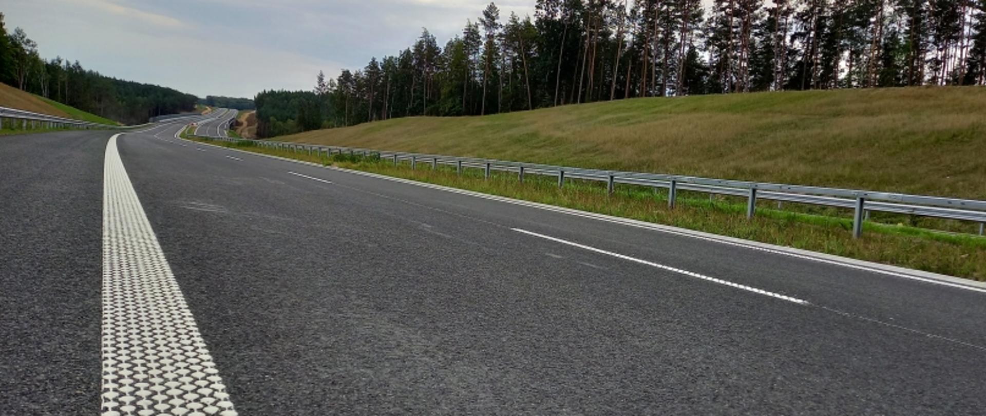 Asfaltowa droga z barierami ochronnymi i zielenią w tle, zakręcająca lekko w lewo.