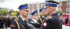 Bliski kadr na oficera Państwowej Straży Pożarnej przypinającego odznaczenie do munduru galowego innego strażaka podczas uroczystego apelu.