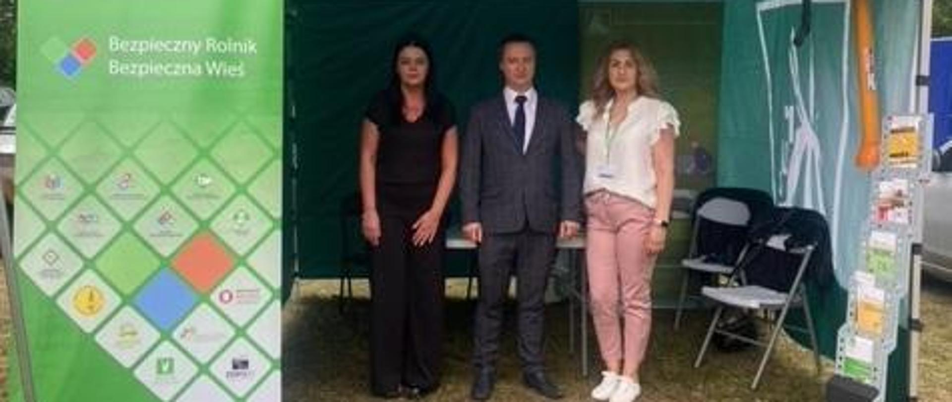 Dwie kobiety i mężczyzna stoją pod namiotem stoiska informacyjnego KRUS, z lewej strony namiotu stoi baner z hasłem: „Bezpieczny Rolnik, Bezpieczna Wieś”
